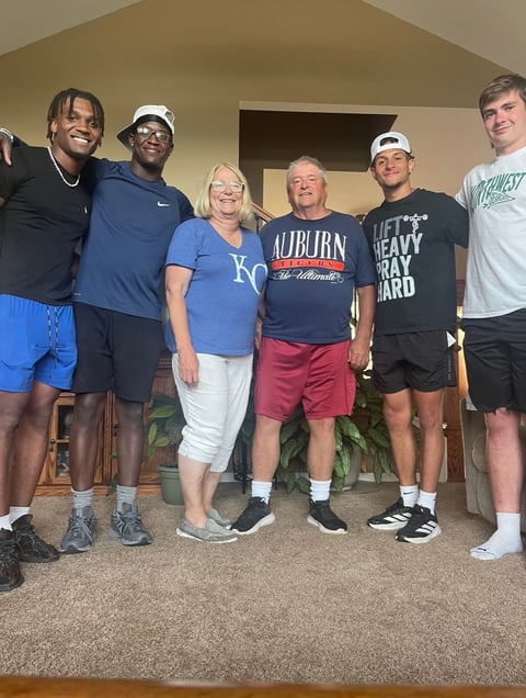 Six people standing together indoors smiling at the camera, wearing casual athletic wear and college team apparel.