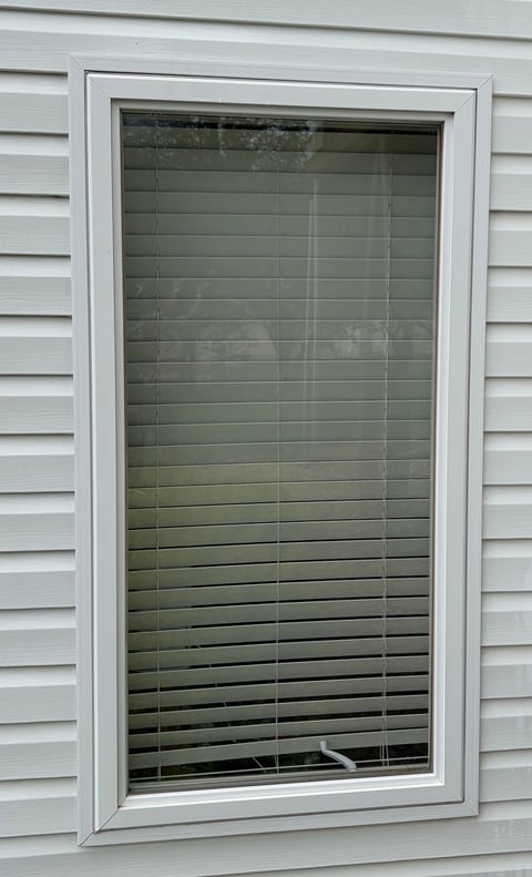 White window frame with closed green horizontal blinds on white vinyl siding