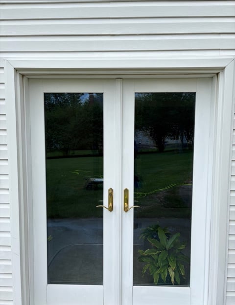 White double French doors with gold handles on white horizontal siding, showing a backyard and green lawn through the glass panes.
