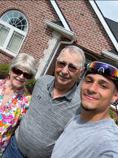 Three people smiling outdoors in front of a brick house on a sunny day